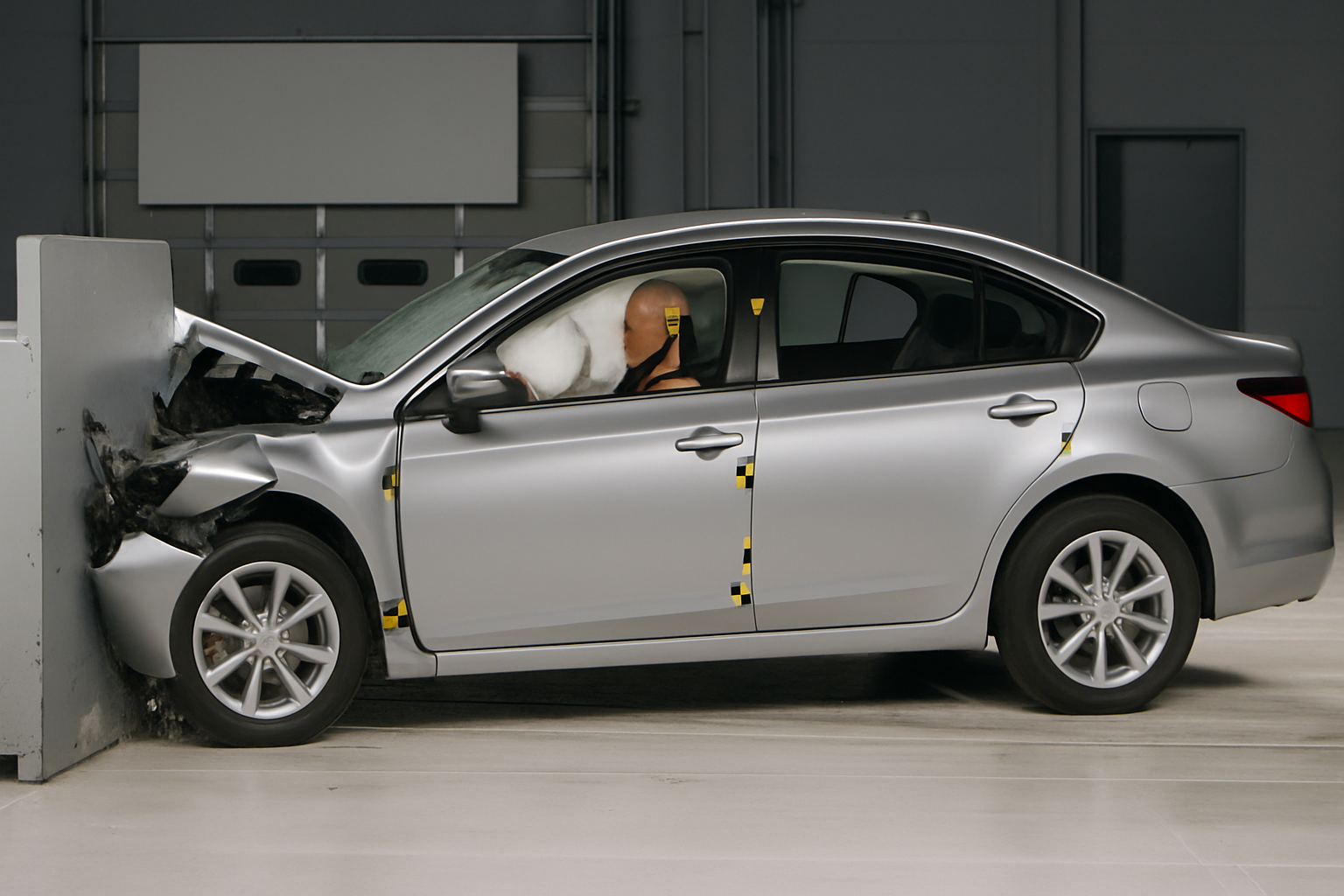 Silver sedan collides with a grey barrier indoors, airbags deployed and a crash test dummy seated inside